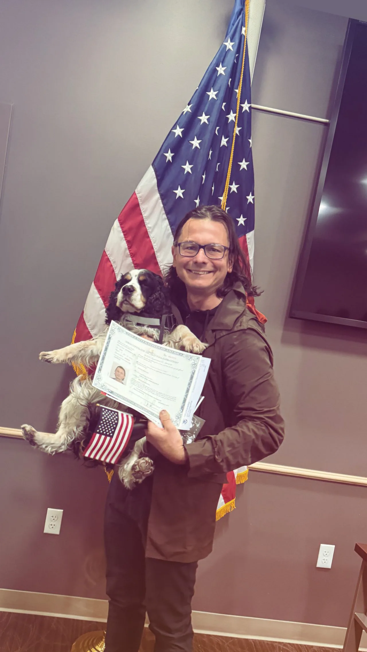 Samuel Andruszkiewicz holding his dog and naturalization certificate in front of an American flag