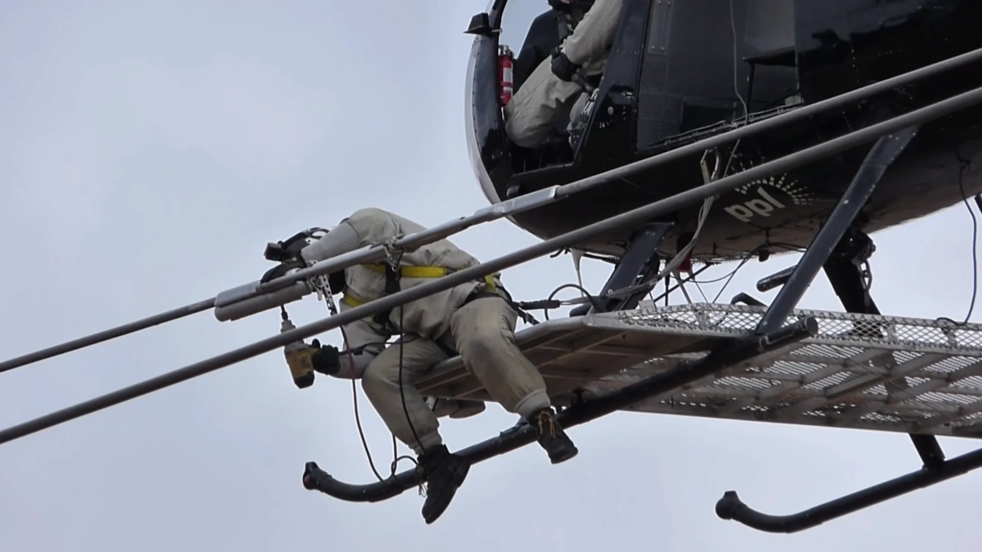 A worker installs equipment on transmission lines from a helicopter platform.