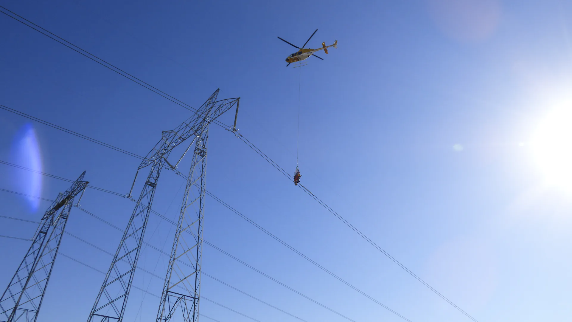 A helicopter suspends an installation crew beside transmission lines and towers.